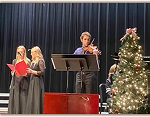 two students singing along with violinist beside Christmas tree